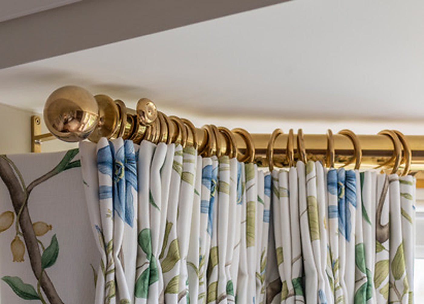 Tillys polished brass bay window curtain pole turning the corner of a bay, with brass rings and floral curtains
